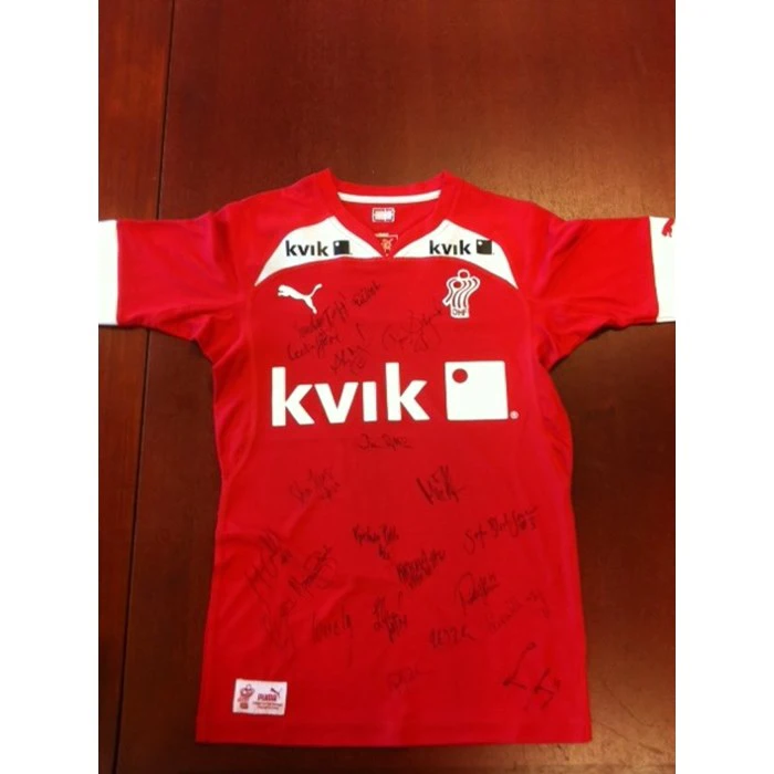 En rød Kvick-sportstrøje med hvide detaljer, Puma-logo og flere underskrifter med sort tusch på forsiden, lagt fladt på et træunderlag.